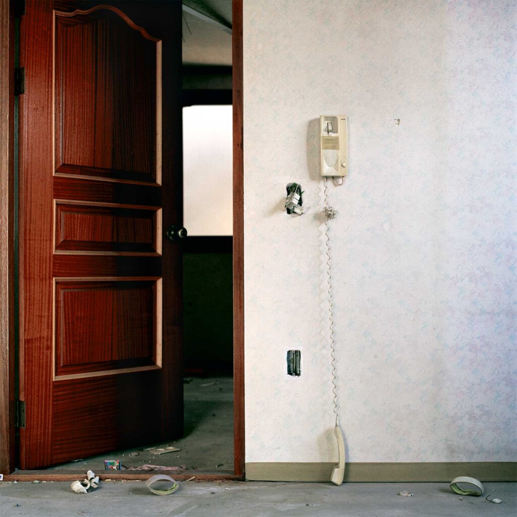 A partially opened wooden door in an abandoned apartment in Seoul, South Korea. Beside the door, a broken intercom telephone hangs off the hook, dangling towards the floor