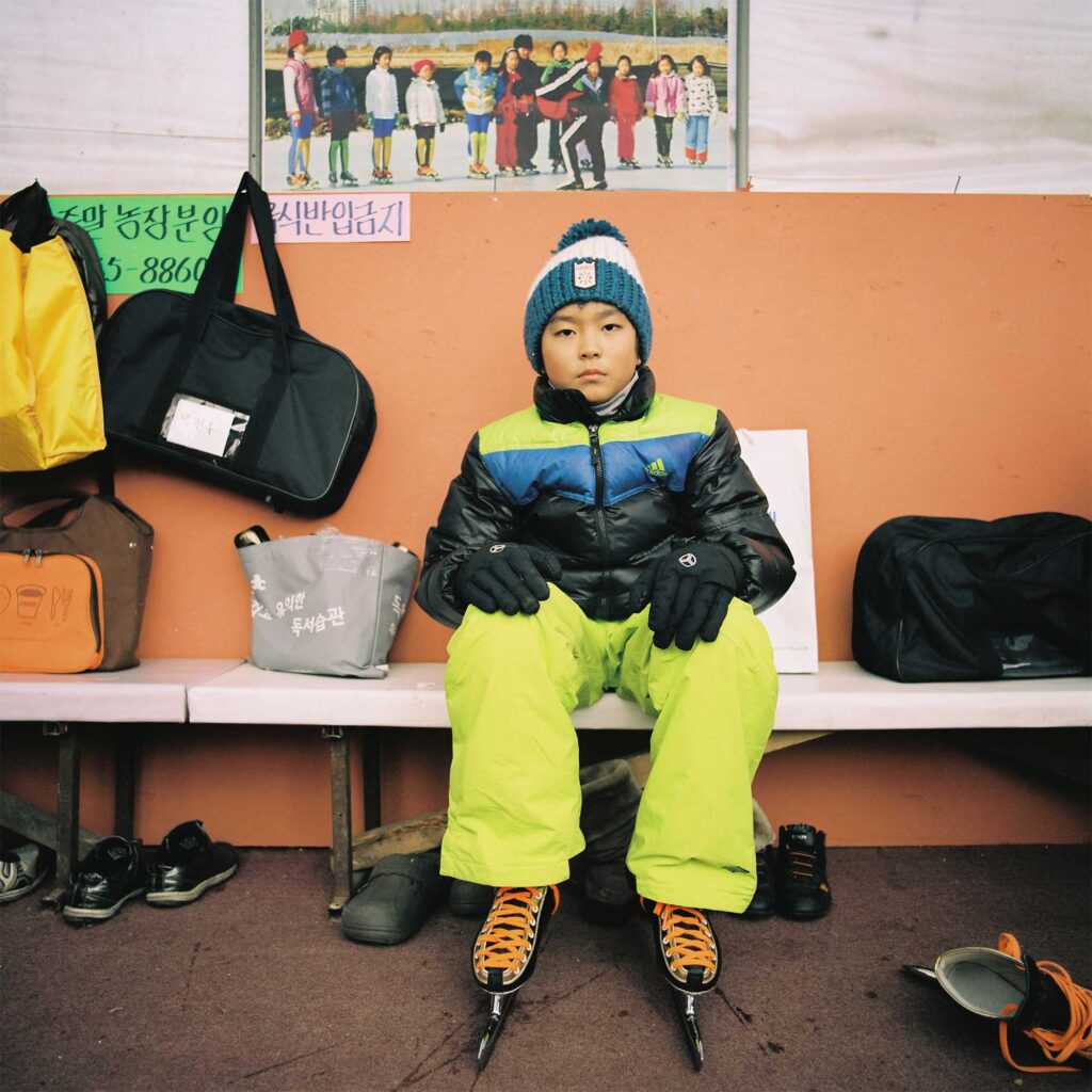 A young Korean boy sits on a bench in the ice rink's changing area, dressed in warm winter clothes and ice skates