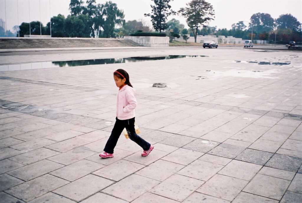 A young girl in black pants and a pink jacket strolls through a spacious, empty square in Pyongyang, North Korea.
