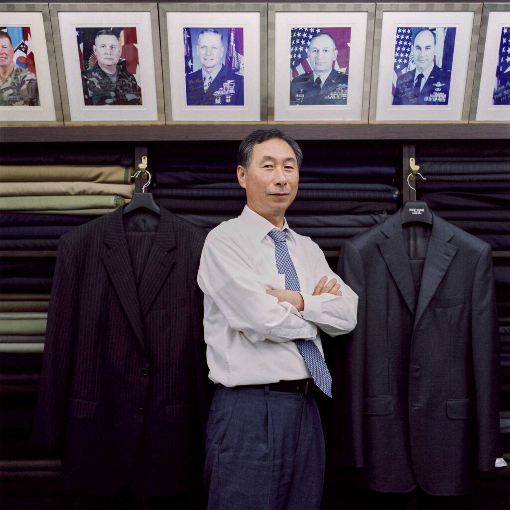 A Korean tailor stands in a shop in Seoul's bustling Itaewon district, surrounded by shelves of vibrant fabrics. Dressed in a white shirt, black pants, and a blue-and-white patterned necktie, he works beneath framed photos of high-ranking American army officers