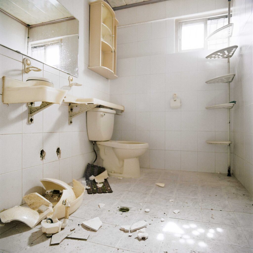 A white bathroom with partially demolished beige-yellowish facilities scattered on the floor. Sunlight streams through the window, casting a dotted pattern on the bathroom floor