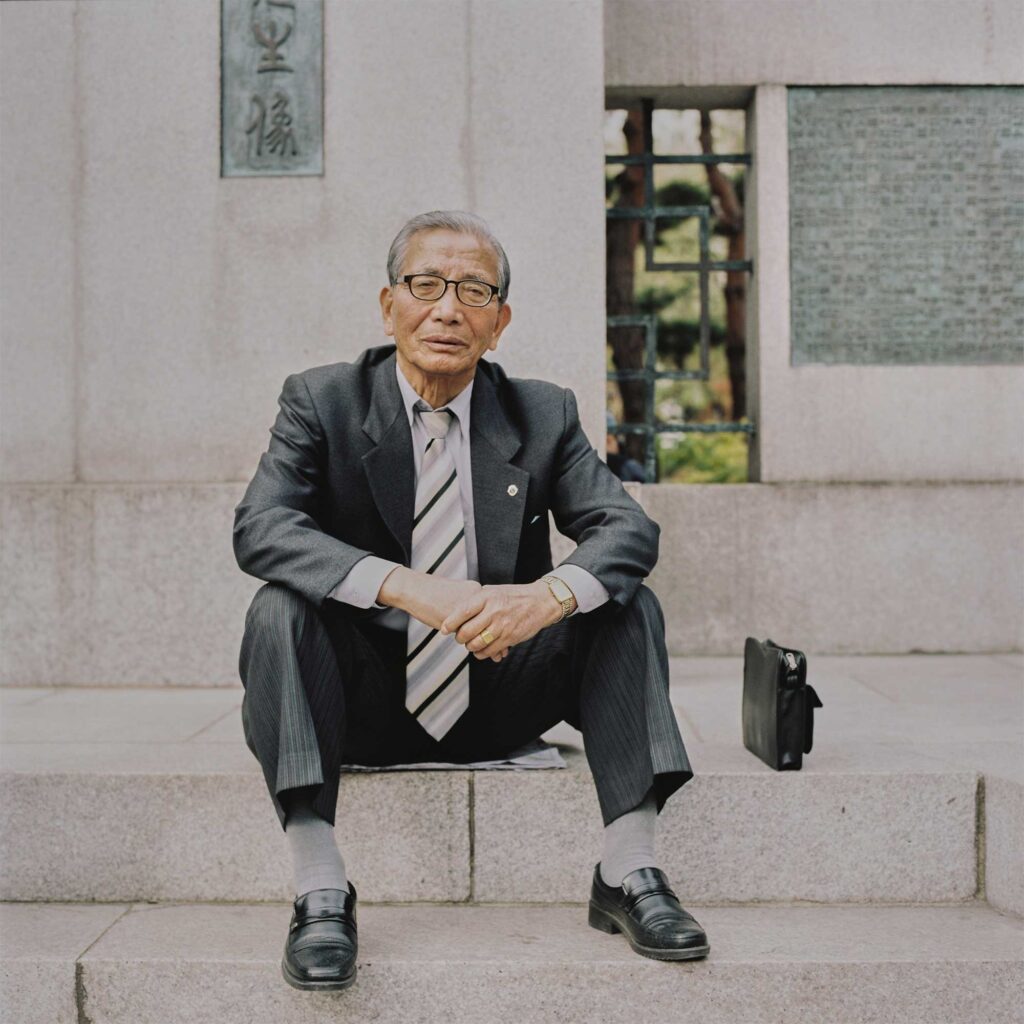 In Seoul, an elderly man sits on a staircase at a park construction site. He wears a grey pinstripe suit, a politely striped necktie, and fashionable glasses, exuding refined elegance