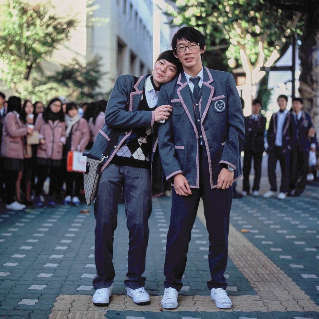 Male high school students in uniform and white sneakers pose, one resting his head on the other's shoulder, amidst a group of classmates