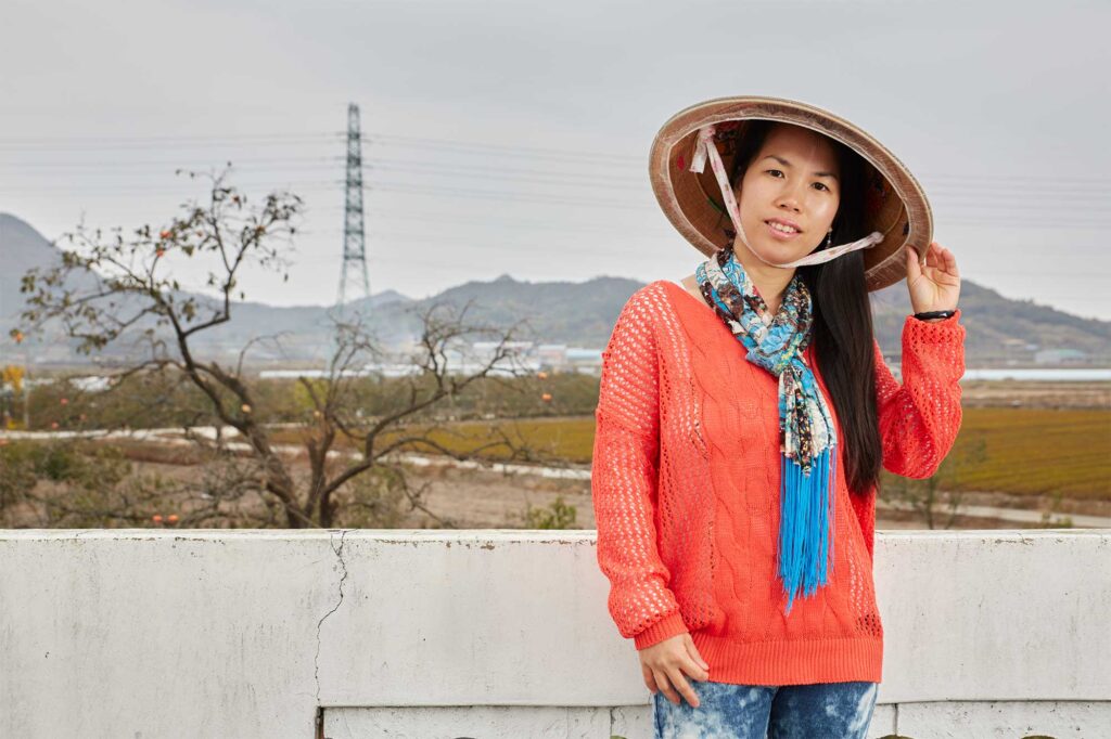 A portrait of a Vietnamese woman in Bongdong, South Korea, wearing an Asian conical hat, a red sweater, blue-dyed jeans, and a scarf