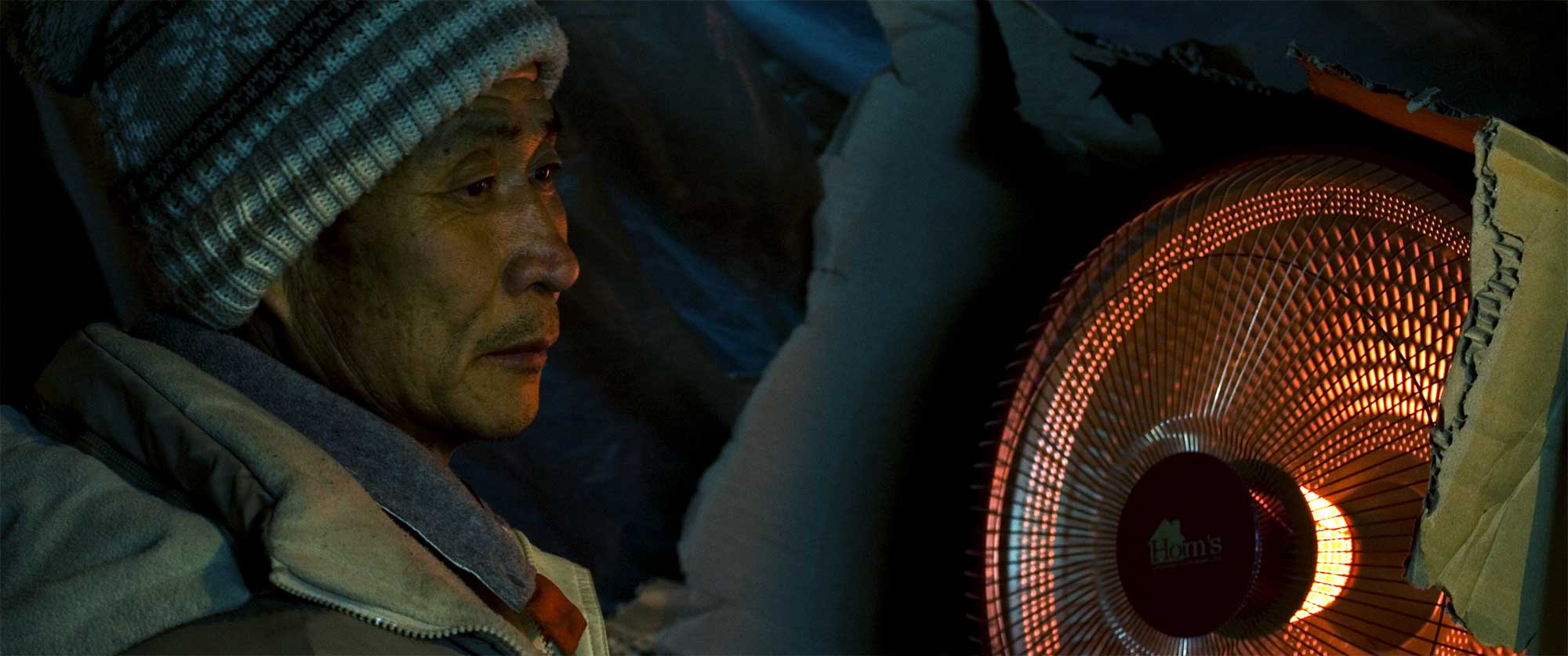 Portrait of a Korean market street vendor embracing the warmth of an electric heater on a winter night