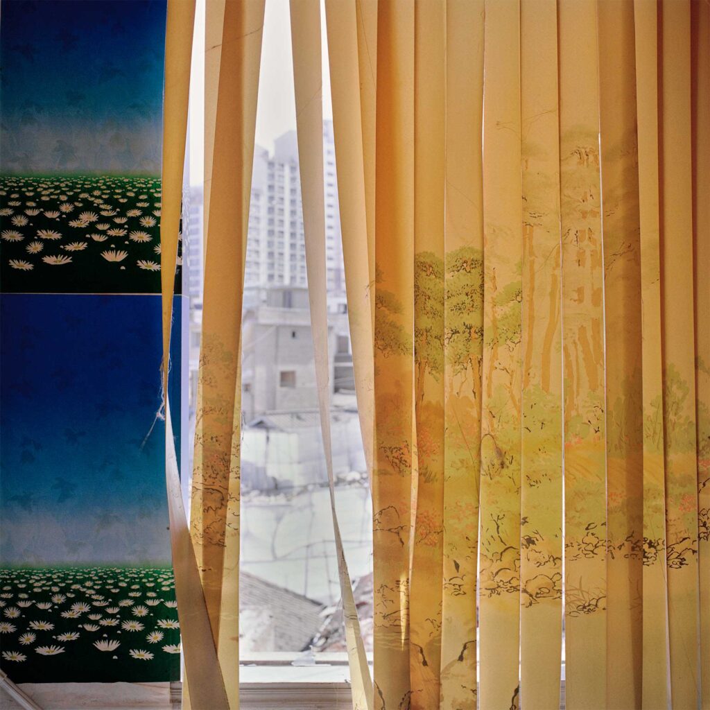 A slat curtain with a forest landscape drawing blows in the wind. Beside the window, a wallpaper depicts daisies against a blue sky. Through the curtain, townhouse buildings in demolition are visible, while newly built apartments rise in Seoul, South Korea