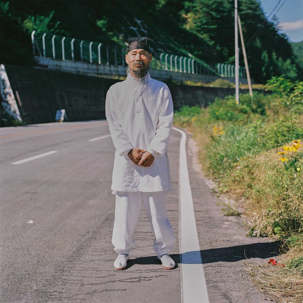 A Korean man in traditional white attire and a black hat, his long gray beard evoking a scholarly aura from ancient times