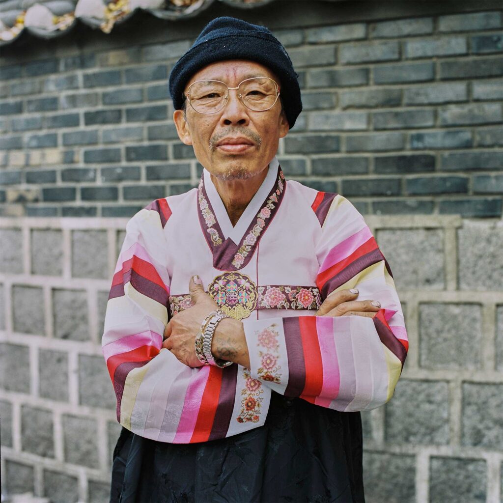 In Seoul, an elderly man stands before a town wall, clad in a colorful Hanbok. His tattooed forearms, beanie hat, and hipster glasses add an unexpected twist to his traditional attire