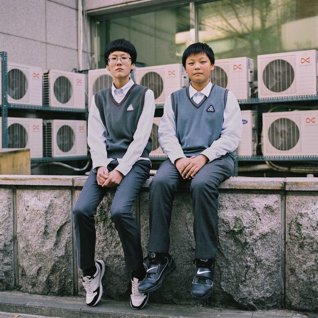 Middle school students sit politely on a wall in front of a building, dressed in grey pants, white shirts, grey sweaters, and sneakers