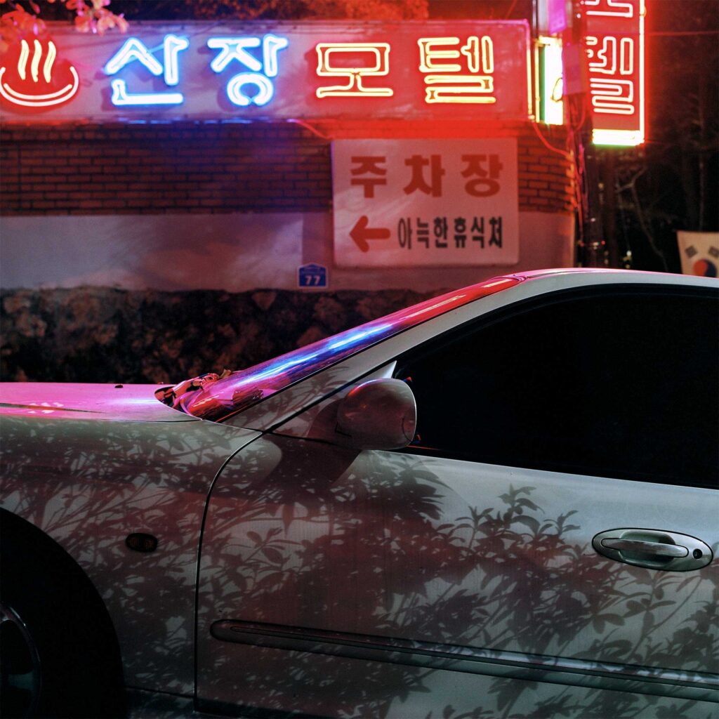 A white car parked at night in front of a sauna building with neon displays outside of Seoul, South Korea