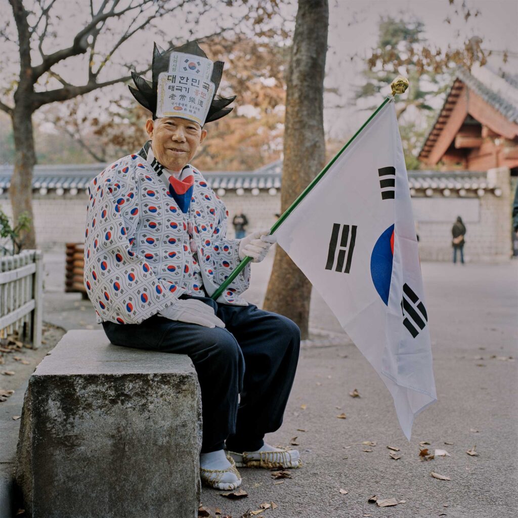 An elderly Korean man sits before a traditional palace in Seoul, clad in traditional attire with a unique twist: a blouse jacket adorned with the Korean Tekukki flag pattern. He holds a large Tekukki flag, smiling at the camera