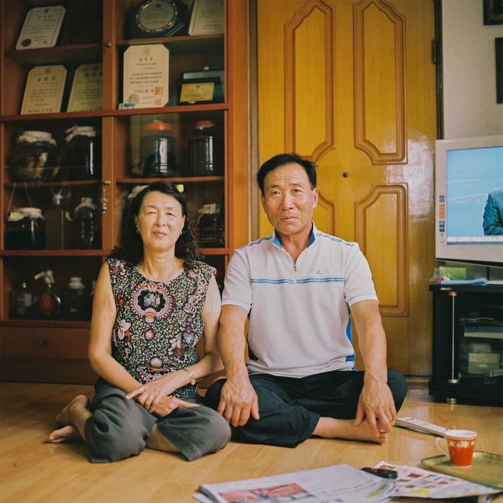 An Korean elderly fishing couple sits on the floor of their living room, surrounded by framed award certificates and pots of homemade traditional Korean liquor.