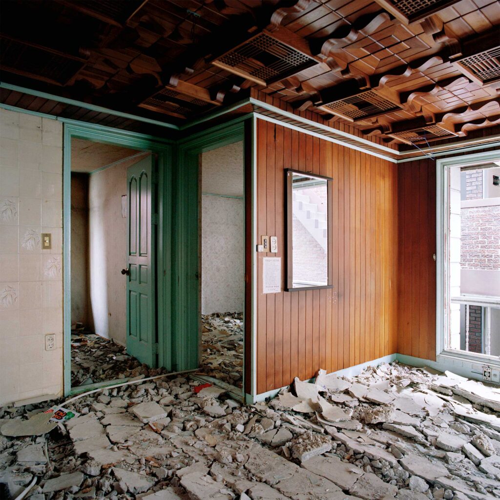 An abandoned living room in an urban redevelopment district of Seoul, South Korea. Wooden panels adorn the walls and ceiling, their ornate patterns contrasting with the demolished concrete floor