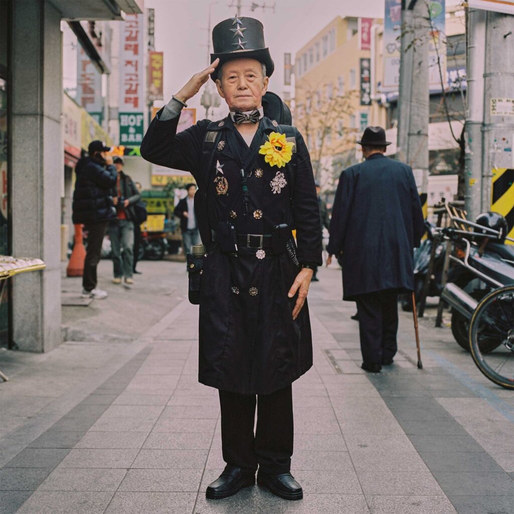 An elderly Korean man, wearing a chimney sweater uniform, salutes the camera on a Seoul sidewalk. Adorned with medals and a large sunflower, his attire exudes character and pride