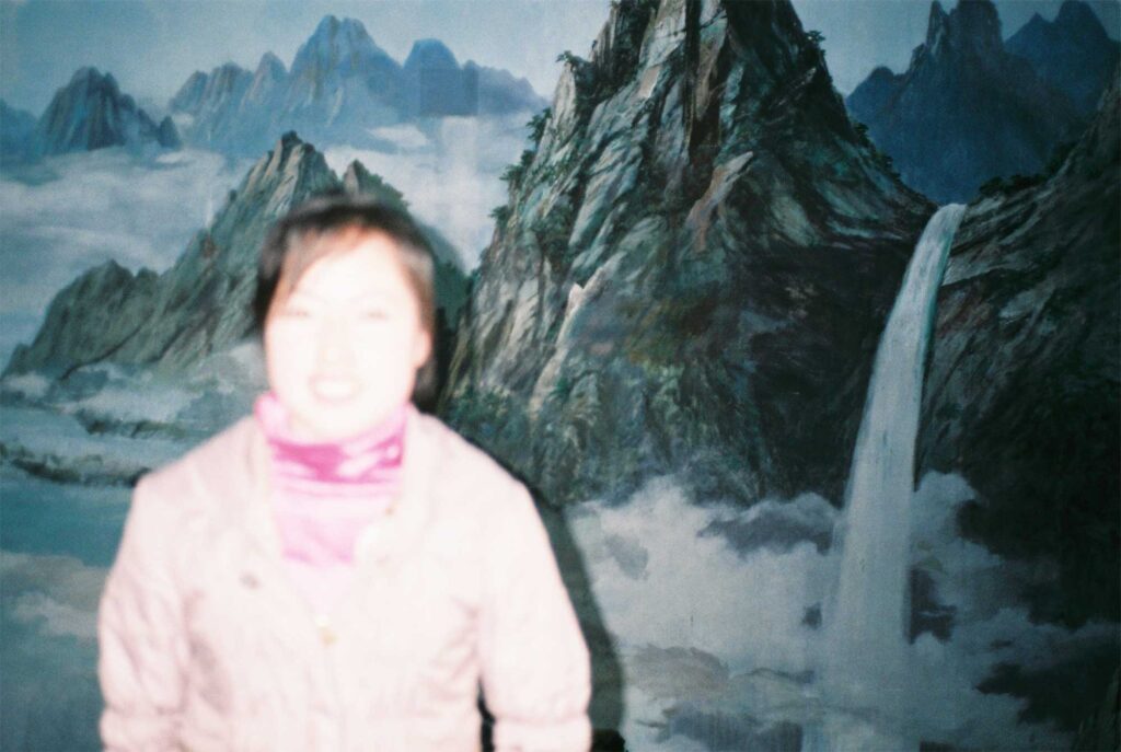 A woman in Pyongyang, North Korea, poses before an interior mural depicting a majestic mountain landscape and waterfall, with the camera focused on the waterfall