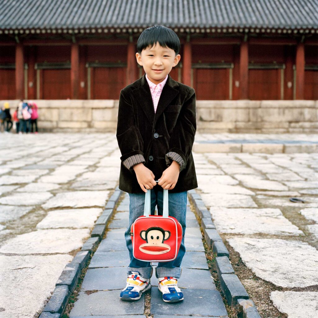 A male Korean primary student standing in front of a traditional Korean palace, holding a bag with a drawing of a monkey face