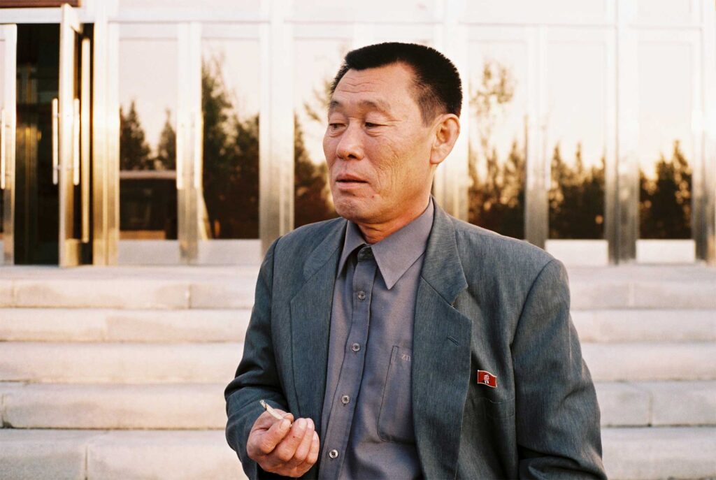 A North Korean tour guide in Pyongyang takes a smoke break in front of the music academy, captured in a portrait