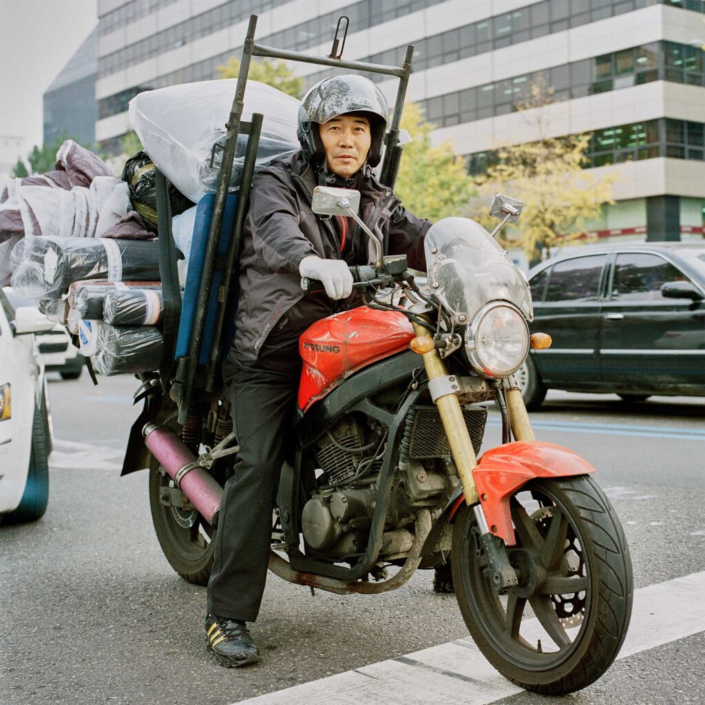 A Korean man sits on his fully loaded delivery bike at a busy road's stoplight, ready to drive off