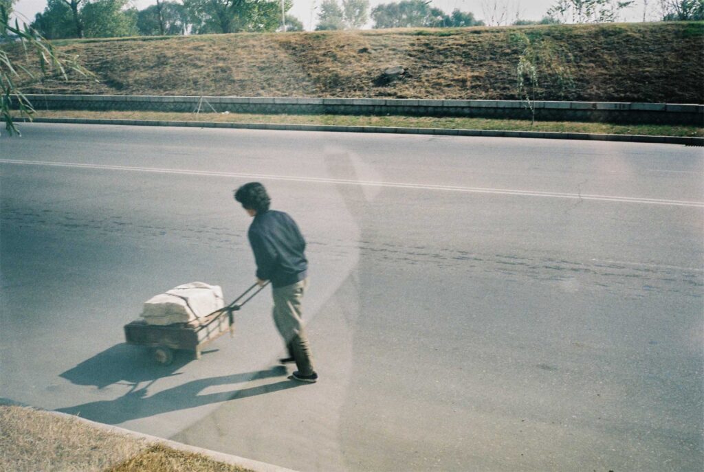 Inside a bus, a snapshot captures a man pushing a heavy cart on a Pyongyang street