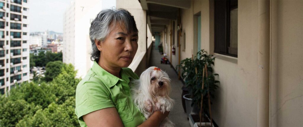 A portrait of a woman holding her small dog in the open hallway of a Seoul apartment building in South Korea