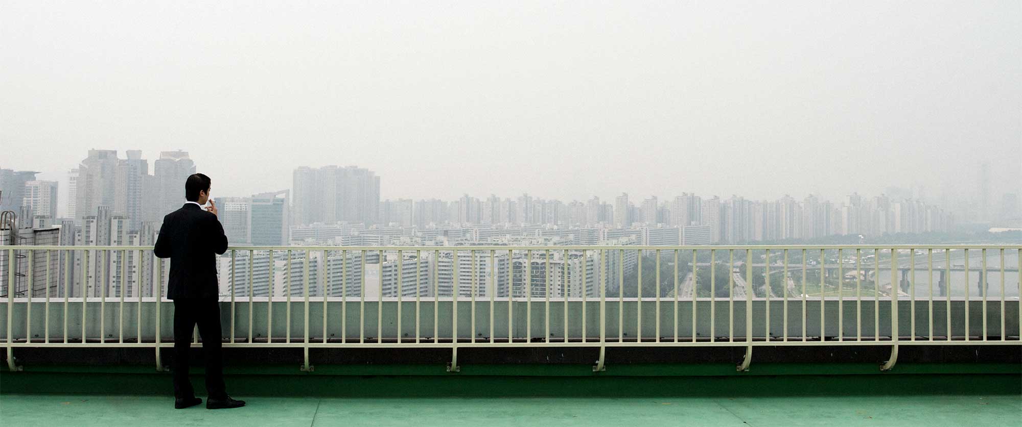 A man dressed in a black suit takes a cigarette break on the helicopter landing platform atop a high-rise building, overlooking the architecture of Gangnam, Seoul, South Korea