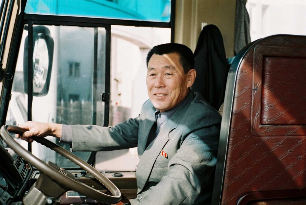 A portrait of a tour bus driver in Pyongyang, North Korea, seated at his driver's seat with his right hand on the steering wheel before takeoff