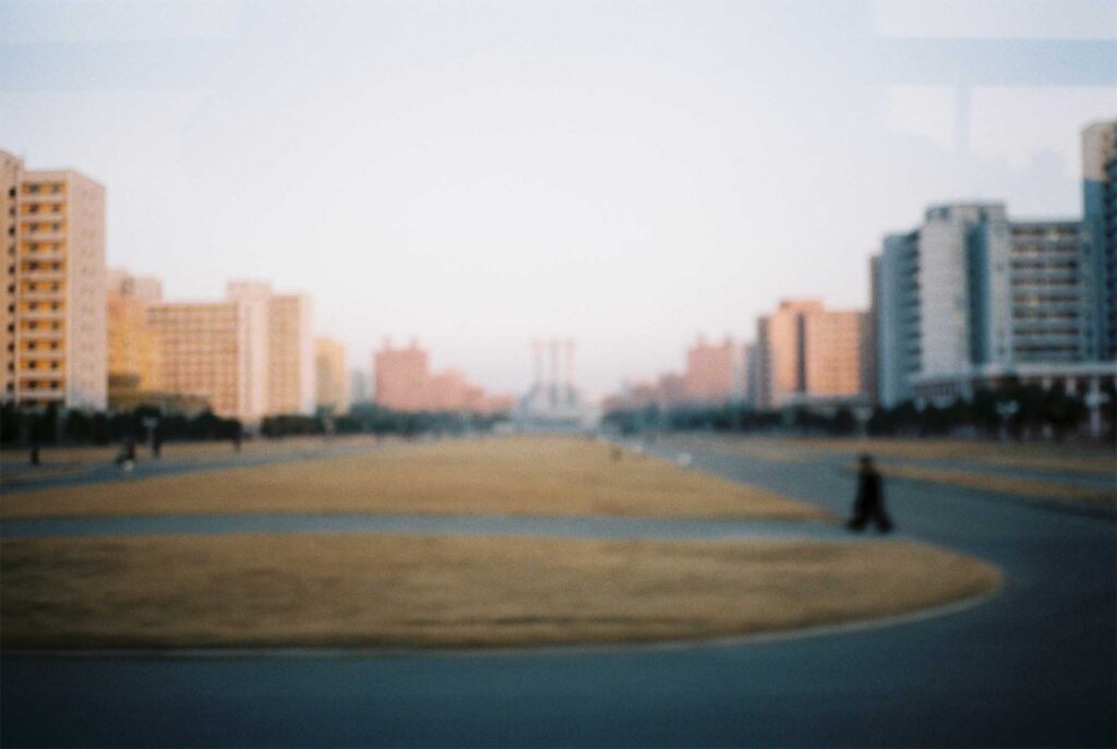 A blurred shot captures a nearly empty promenade in Pyongyang, North Korea, flanked by apartment buildings and leading towards a prominent monument
