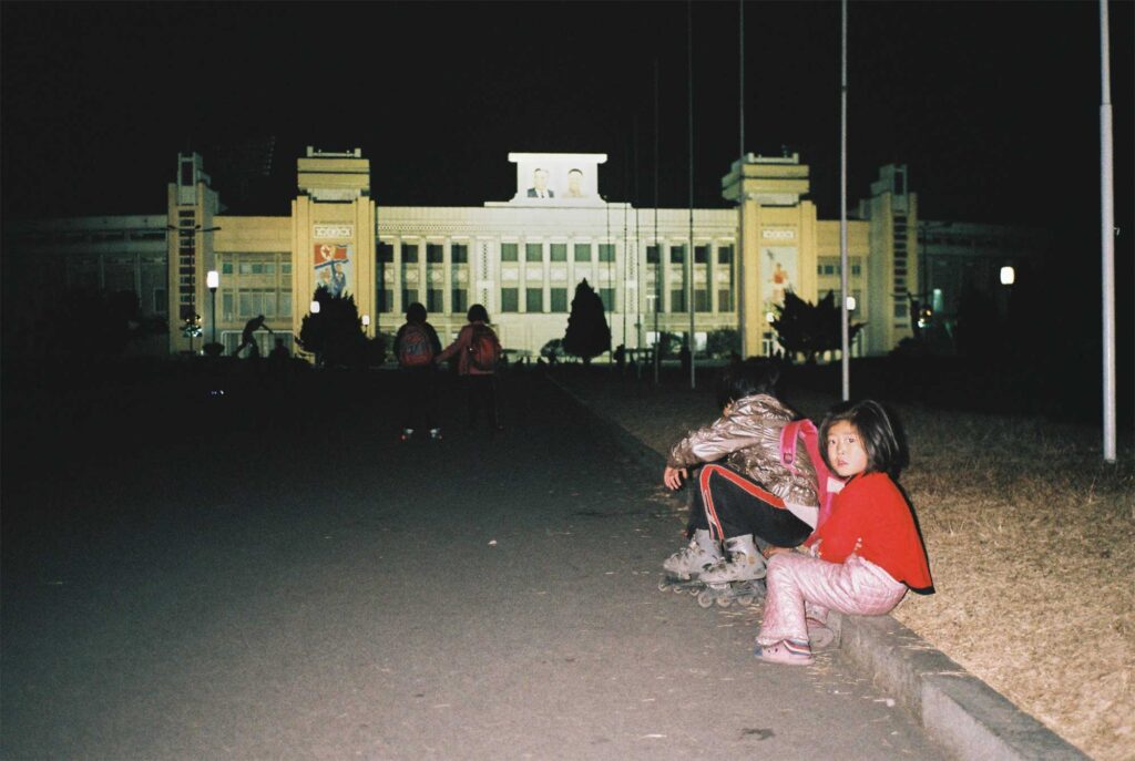 Children in Pyongyang, North Korea, play on roller blades in the dark, with a monumental building in the background adorned with images of the late Korean leaders Kim Jong-il and Kim Il-sung
