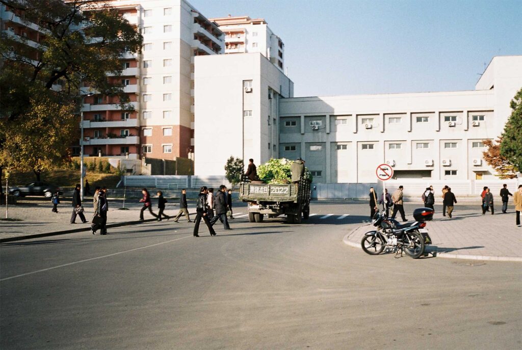 A street corner in Pyongyang, North Korea, with apartment buildings in the background, pedestrians crossing the street, and a truck loaded with Kimchi cabbages parked