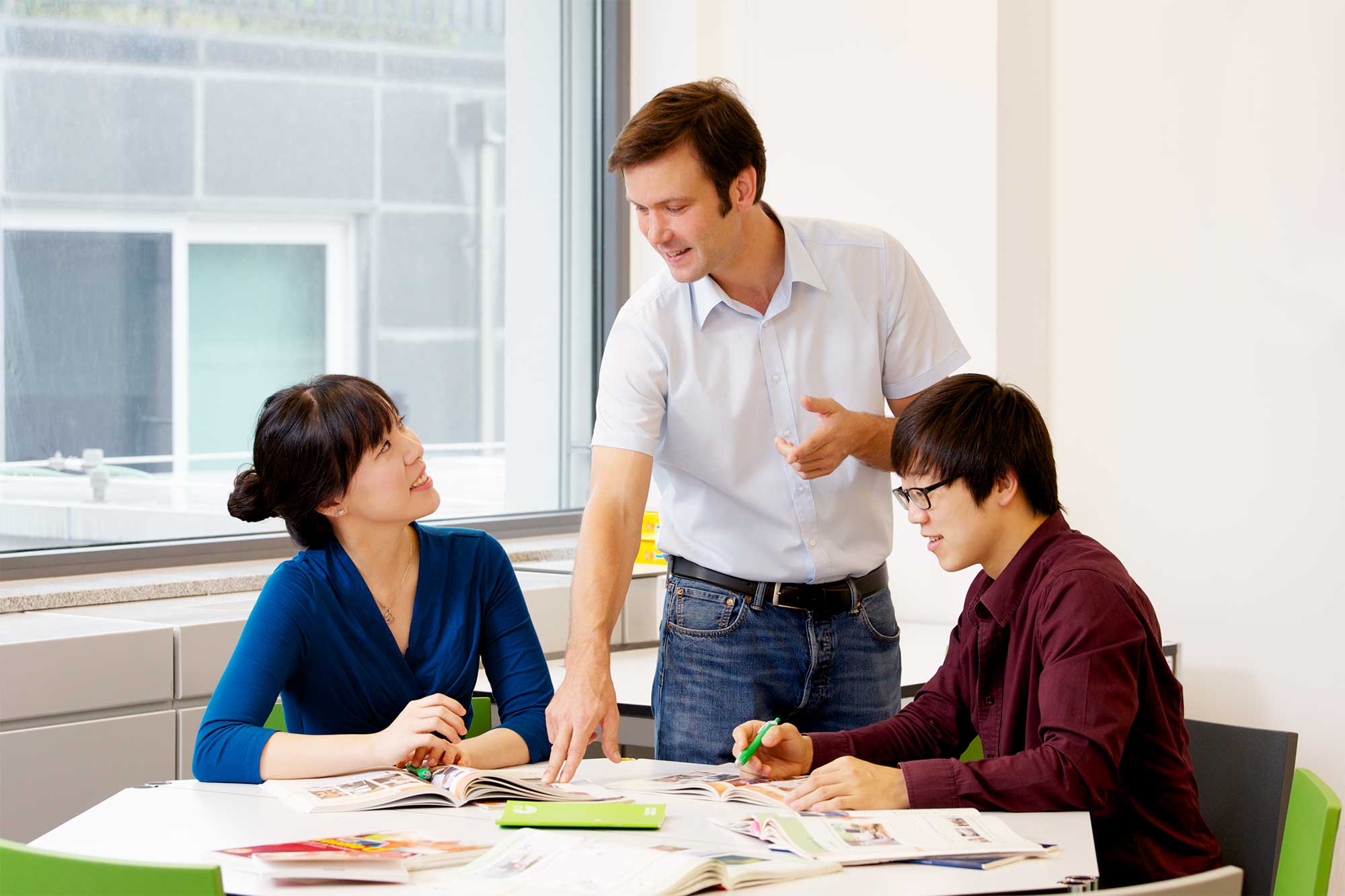 A young male teacher at the Goethe Institut in Seoul assists two Korean students with classroom assignments aimed at improving their German language skills.