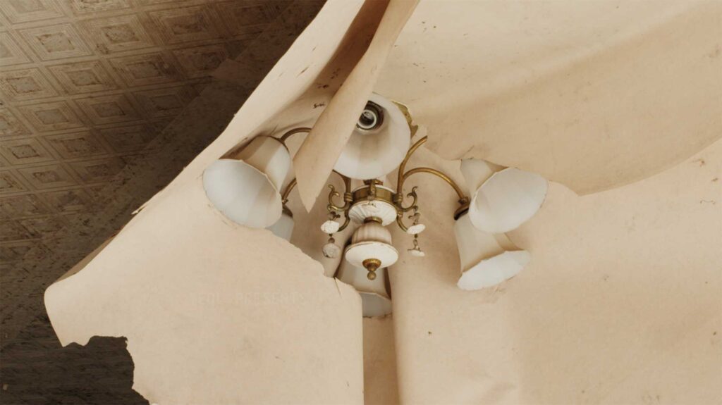 Wallpaper peeling from the ceiling, wrapped around a chandelier in the living room featured in the film Translating Furniture, showcasing Kwangho Lee's award-winning work.