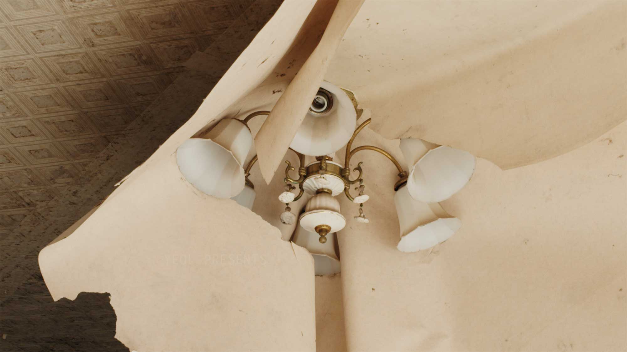 Wallpaper peeling from the ceiling, wrapped around a chandelier in the living room featured in the film Translating Furniture, showcasing Kwangho Lee's award-winning work.