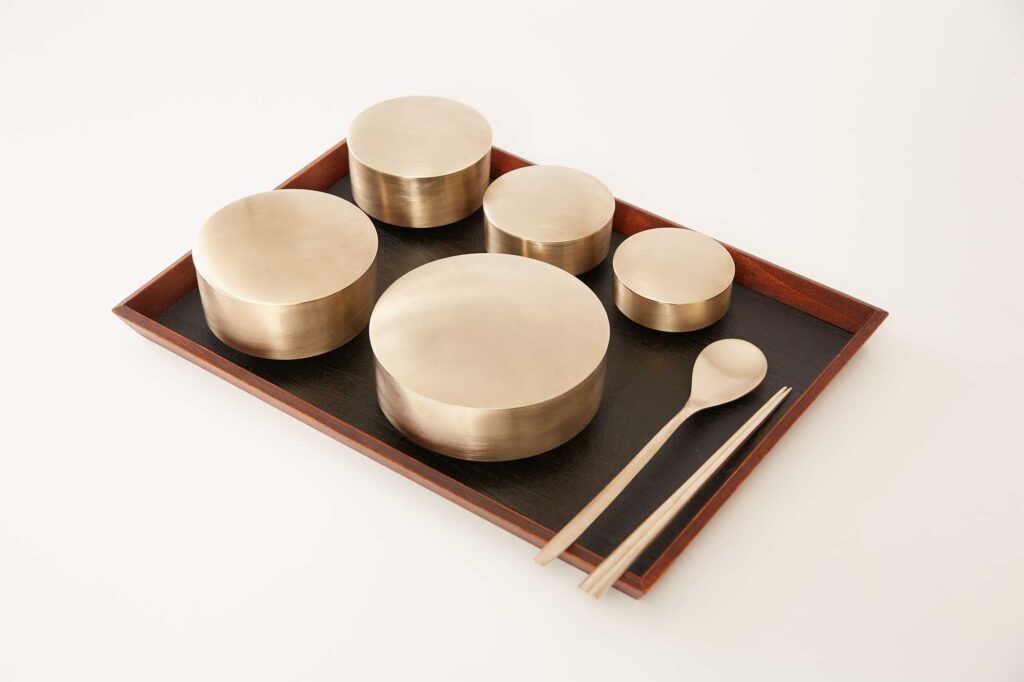A tablet displays Korean traditional brass kitchenware, including a soup bowl, rice bowl, chopsticks, and spoon designed by Gio Kisang and crafted by YÉOL Artisan of the Year, Kim Soo-Young.