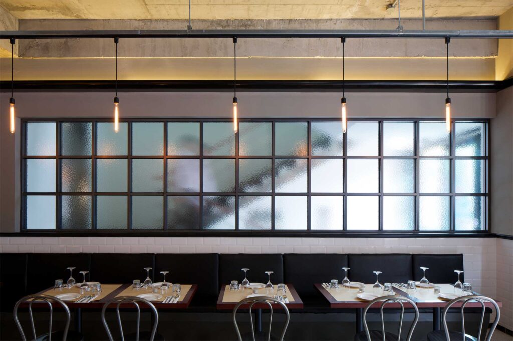 Row of tables and chairs in front of a long seating area with a milky glass window, part of Libertine bar and restaurant in Itaewon, Seoul, with a modern, minimalist design.