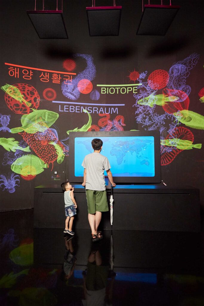 A father and son stand in front of a large digital world map display in an ocean-themed exhibition space at the German Pavilion during Germany’s National Day at the World Exhibition in Yeosu, South Korea.