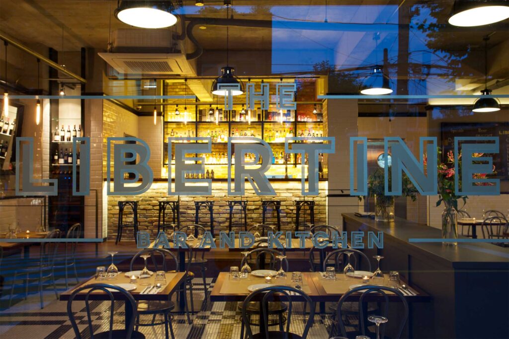 An exterior shot of the logo on the large glass front of Libertine bar and restaurant in Itaewon, Seoul, revealing a modern, minimalist design with warm, ambient lighting inside.
