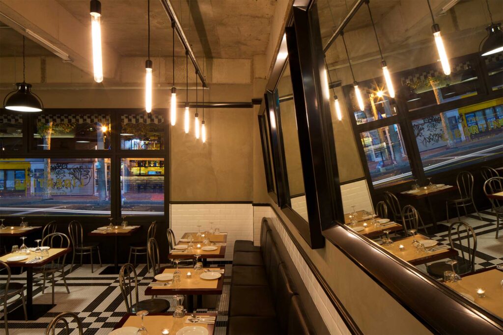 Big mirrors create depth above a row of tables and chairs along a long seating area in Libertine bar and restaurant in Itaewon, Seoul, with a modern, minimalist design.