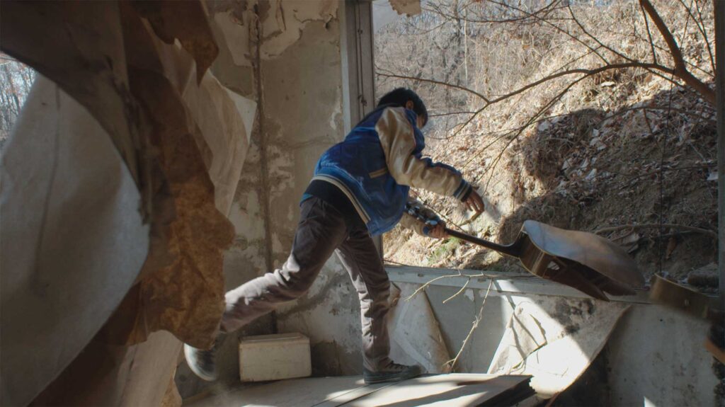 A boy explores an abandoned building, smashing a guitar onto the window sill, featured in the film Translating Furniture, showcasing Kwangho Lee's award-winning work.