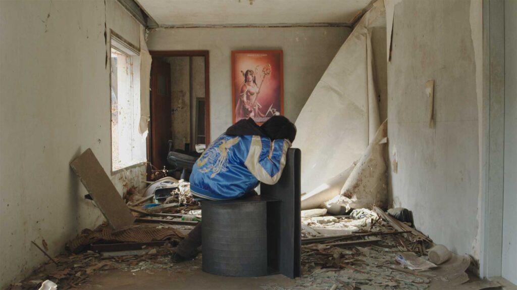 A boy explores an abandoned building, drawn to a framed poster of a goddess-like woman on the wall, featured in the film Translating Furniture, showcasing Kwangho Lee's award-winning work.