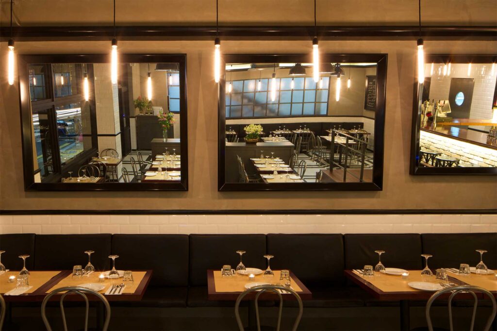 Evening interior with a row of tables and chairs in front of a long seating area with a milky glass window, part of Libertine bar and restaurant in Itaewon, Seoul, featuring a modern, minimalist design with ambient lighting.