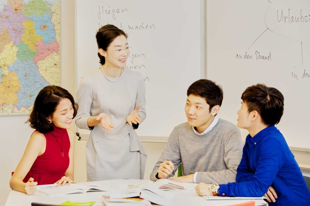 A female Korean German language teacher at the Goethe Institut in Seoul assists three Korean students with classroom assignments to improve their German language skills.
