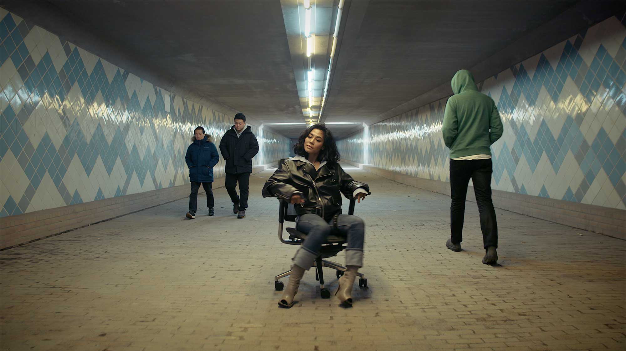 A young woman sits on an office chair in an underground passage as passersby walk past her at night in Seoul, South Korea.