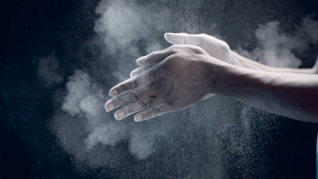 Hands covered in chalk clap, sending white particles flying through the air, creating a striking contrast against a black background for a Millet sports fashion campaign.