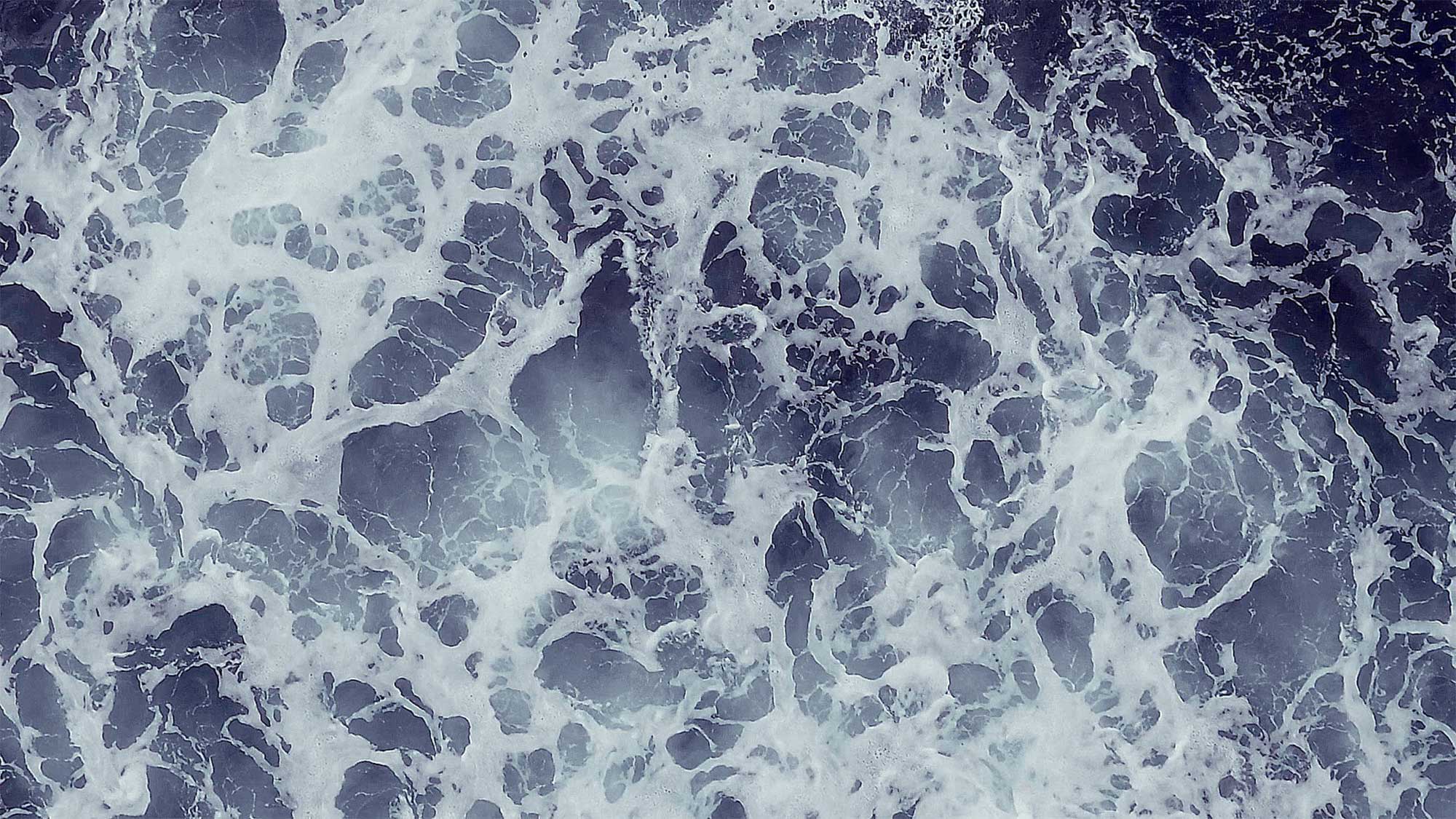 The crashing waves swirl together in shades of blue and white, creating a dynamic, almost painterly texture on the ocean’s surface.