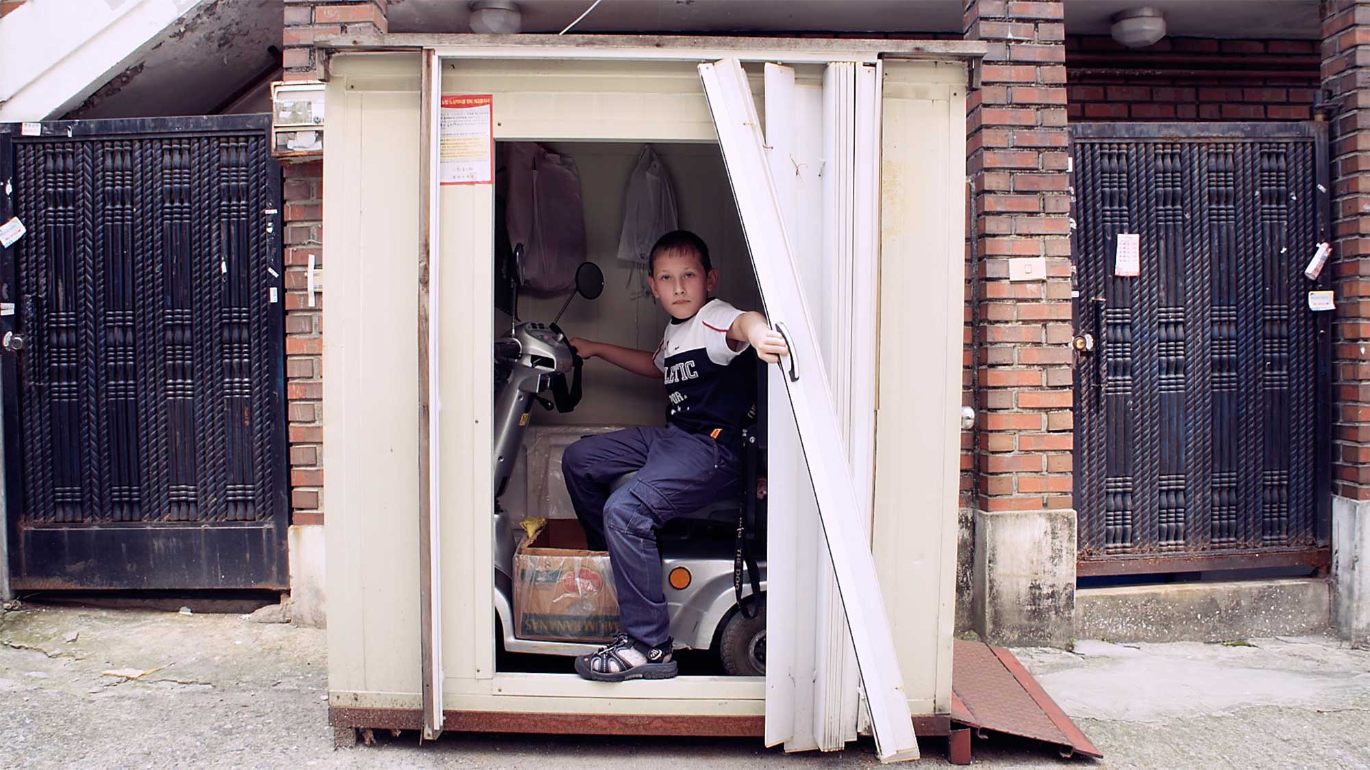 Young boy sits in storage space on handicap vehicle in old run-down neighborhood in Seoul, South Korea, pulling open curtain and posing to camera.