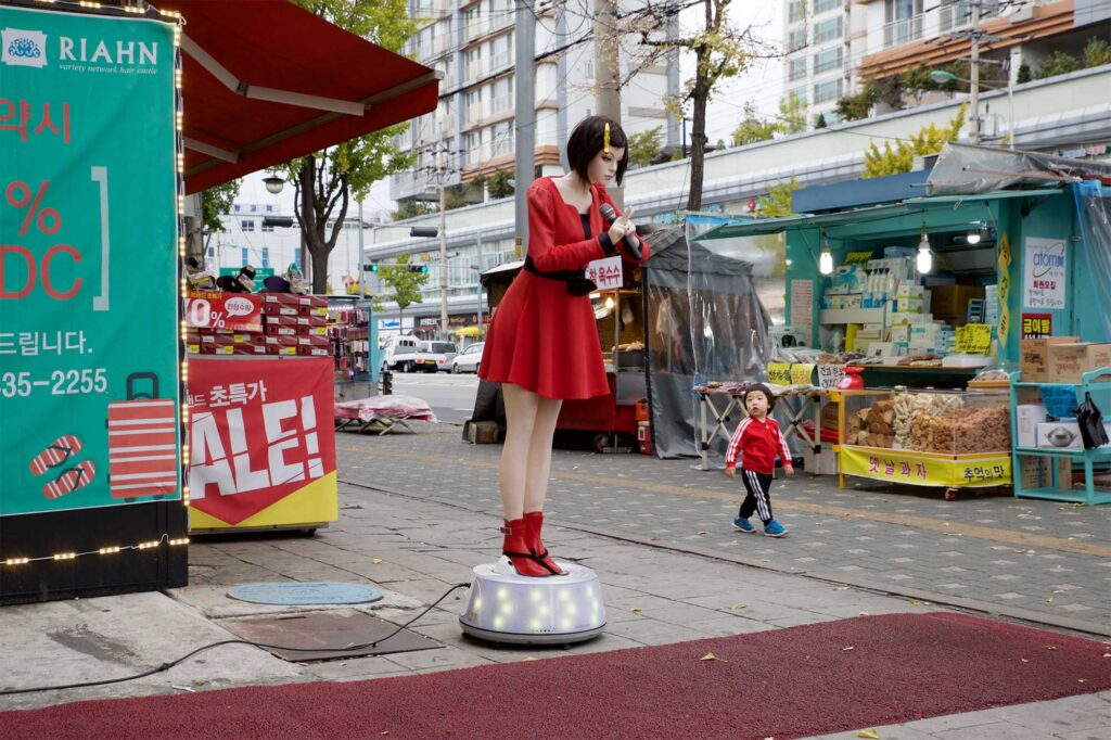A human-like plastic doll stands outside a hairdressing salon in Seoul, South Korea, dressed in red and black with a yellow peg in her hair, catching the attention of a small boy walking by.