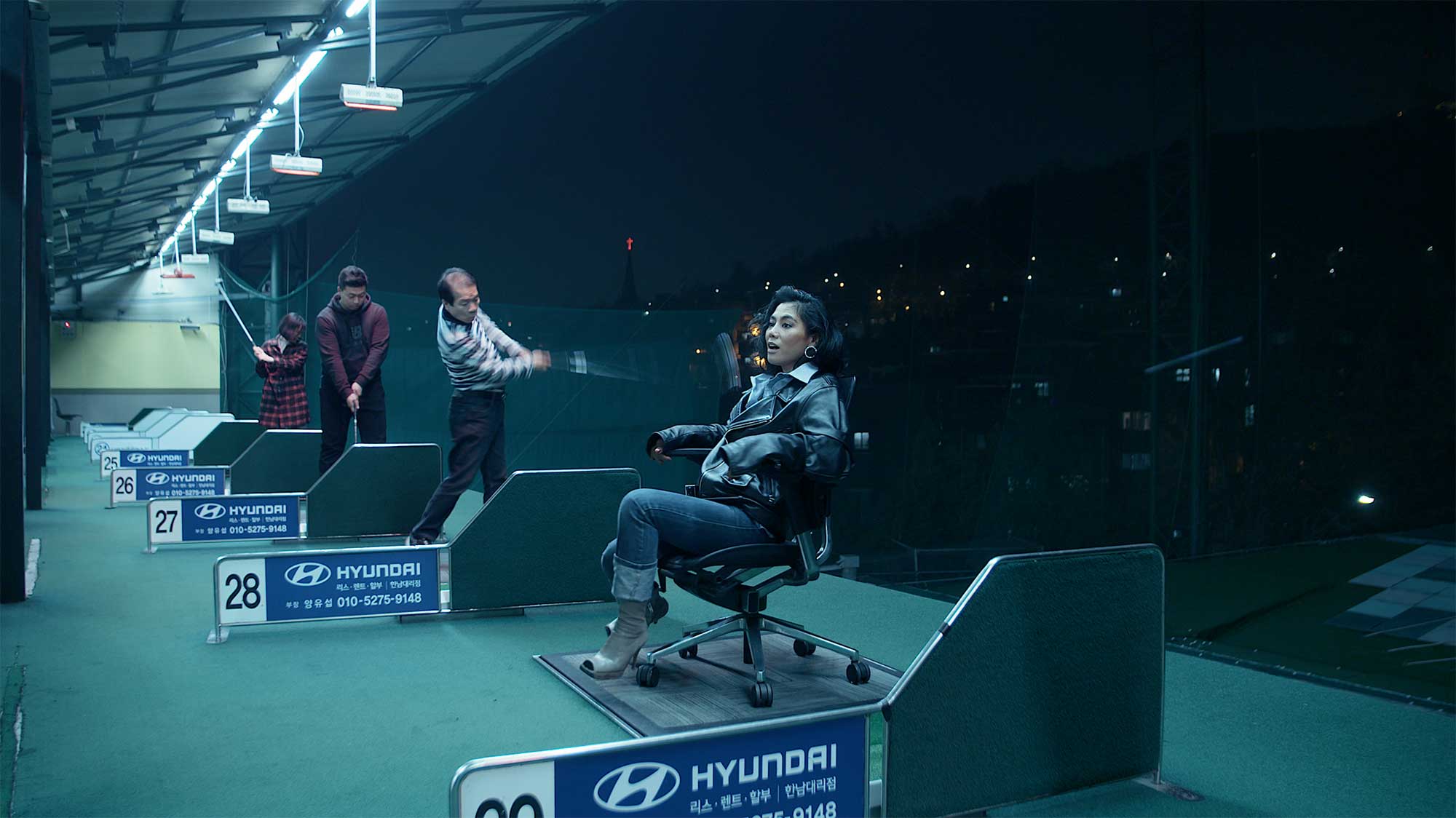 A young woman sits on an office chair at the tee-off area of a golf driving range as golfers hit balls into the distance at night in Seoul, South Korea.