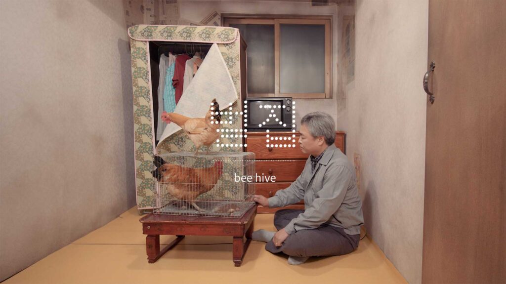 An aged former factory worker sits on the floor beside a small table with a chicken cage, one chicken inside and one outside, in his cramped bee hive home from Nils Clauss's award-winning short film Bikini Words.