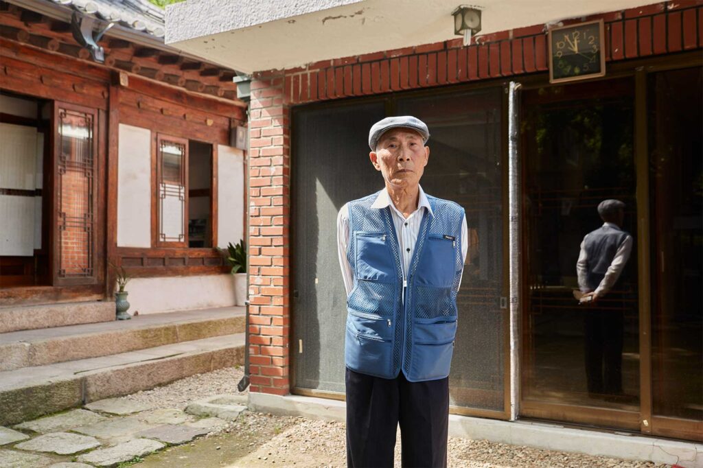 Portrait of a Korean man in the courtyard of his home, blending traditional Korean architecture with Western-style brick elements in Seoul, South Korea.