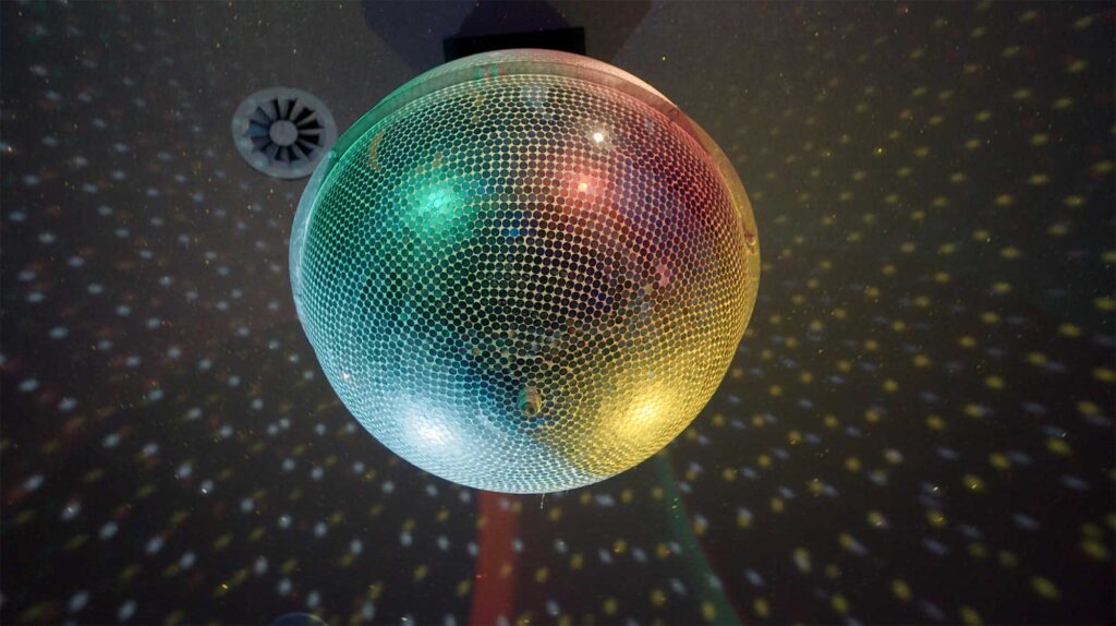 A large mirror ball rotates under shifting colored lights, hanging from the ceiling of an old-style Korean dance hall, from Nils Clauss's award-winning short film Bikini Words.